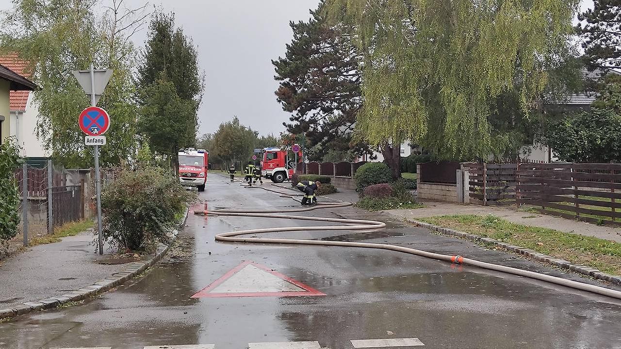 Eine Ortsstraße an einem bewölkten Tag. Der Boden ist nass, im Hintergrund sind zwei Feuerwehrautos zu sehen. Auf der Straße liegt ein Schlauch.