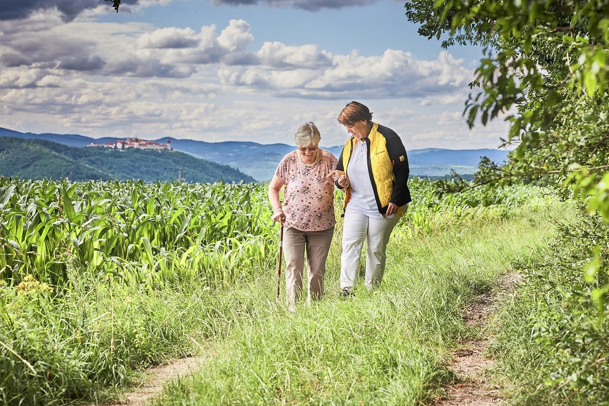 Zwei Personen stehen in der Natur, im Hintergrund sieht man den Himmel. Eine jüngere Person hält die Hand einer älteren Person, während die ältere Person versucht, mit dem Stock den nächsten Schritt zu machen.