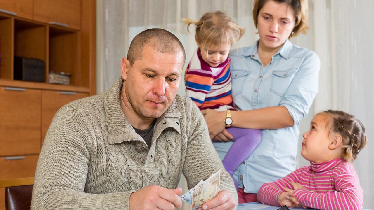 Ein Vater sitzt am Tisch und zählt die Banknoten, während die Mutter mit zwei Kindern im Hintergrund zusieht.