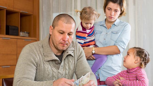 Ein Vater sitzt am Tisch und zählt die Banknoten, während die Mutter mit zwei Kindern im Hintergrund zusieht.