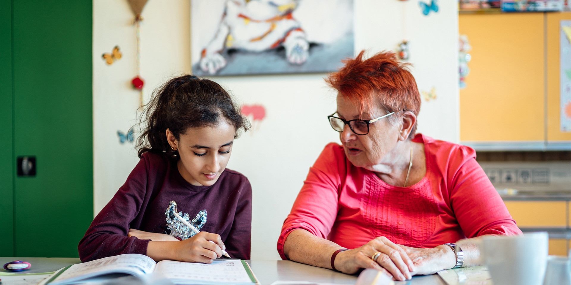 Ein etwa 10-jähriges Mädchen mit dunklen Haaren und eine ältere Dame mit rotgefärbten Haaren sitzen nebeneinander und blicken auf dasselbe Schulbuch. Im Hintergrund sind bunte Kästen und Bilder zu sehen, im Vordergrund liegen Schnellhefter und ein Notizbuch.