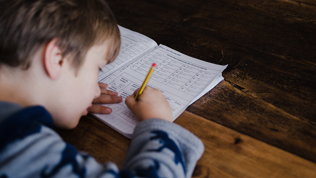 Auf dem Bild ist ein Bub mit blauen Pullover zu erkennen. Das Kind beugt sich nach vorne über ein Blatt Papier und hält einen Stift in der Hand. 