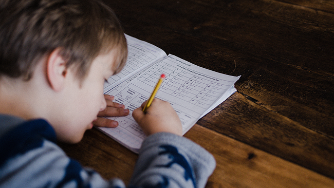 Auf dem Bild ist ein Bub mit blauen Pullover zu erkennen. Das Kind beugt sich nach vorne über ein Blatt Papier und hält einen Stift in der Hand. 