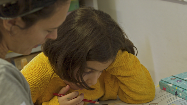 Auf dem Bild ist ein Mädchen mit einem gelben Pullover und dunklen kurzen Haaren abgebildet. Das Mädchen lernt. Eine Frau vom Roten Kreuz hilft beim Lernen und erklärt.