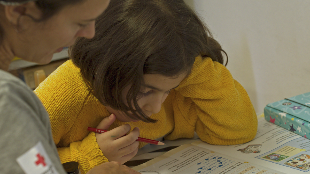 Auf dem Bild ist ein Mädchen mit einem gelben Pullover und dunklen kurzen Haaren abgebildet. Das Mädchen lernt. Eine Frau vom Roten Kreuz hilft beim Lernen und erklärt.