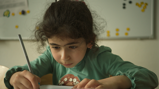 Auf dem Bild ist ein kleines Mädchen mit dunklen Haaren und einem langen türkisen T-Shirt zu sehen. Das Mädchen hält einen Stift in der rechten Hand und blickt auf ein Blatt Papier. 