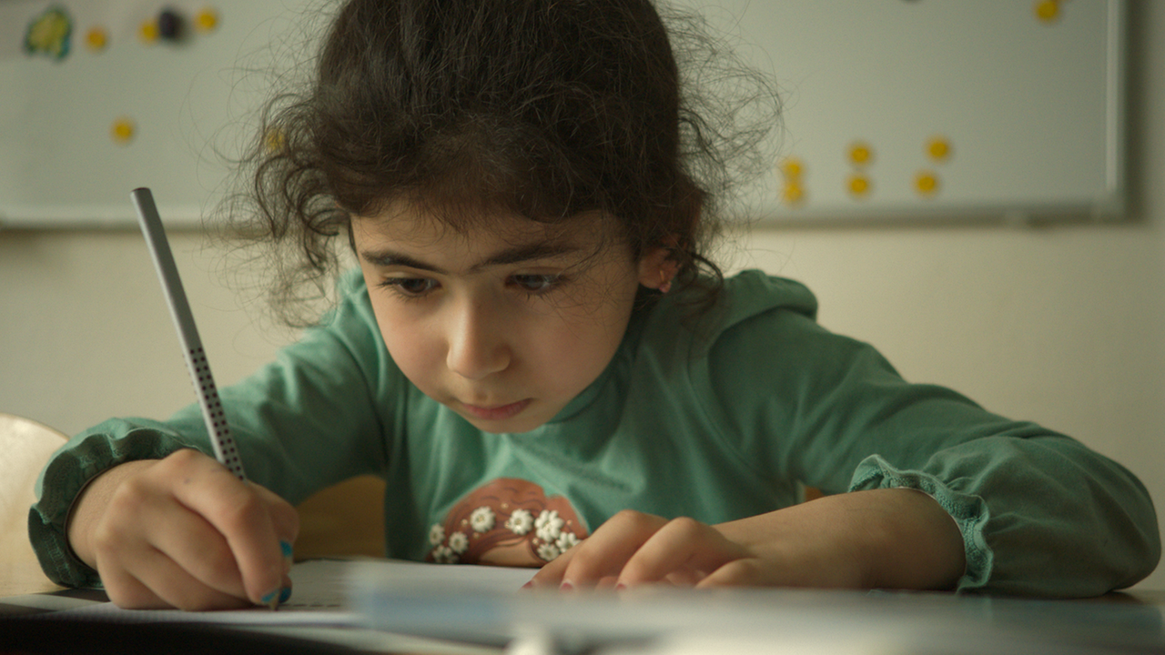 Auf dem Bild ist ein kleines Mädchen mit dunklen Haaren und einem langen türkisen T-Shirt zu sehen. Das Mädchen hält einen Stift in der rechten Hand und blickt auf ein Blatt Papier. 