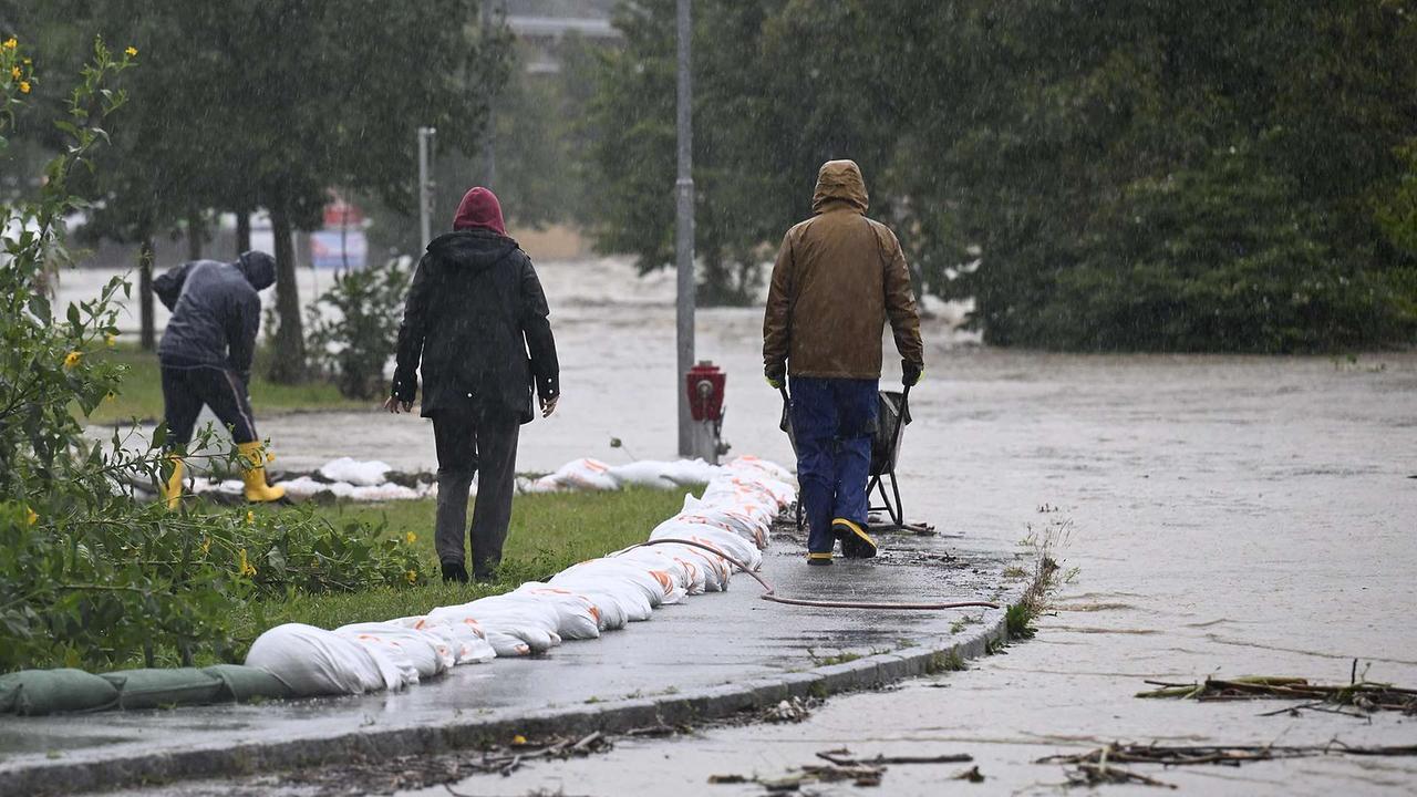 Eine überschwemmte Straße. Es sind Menschen zu sehen, die Sandsäcke entlang des Gehsteiges verteilen.