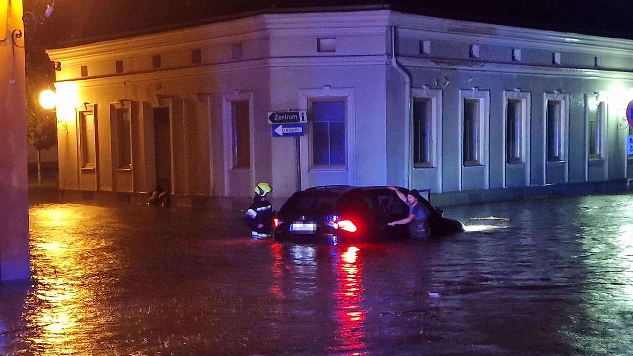 Nacht: Ein dunkler PKW steht bis zur Nummerntafel unter Wasser. Jemand in Feuerwehruniform und ein Mann in Zivilkleidung stehen daneben, beide sind bis zur Hüfte im Wasser. Im Hintergrund ein Gebäude.