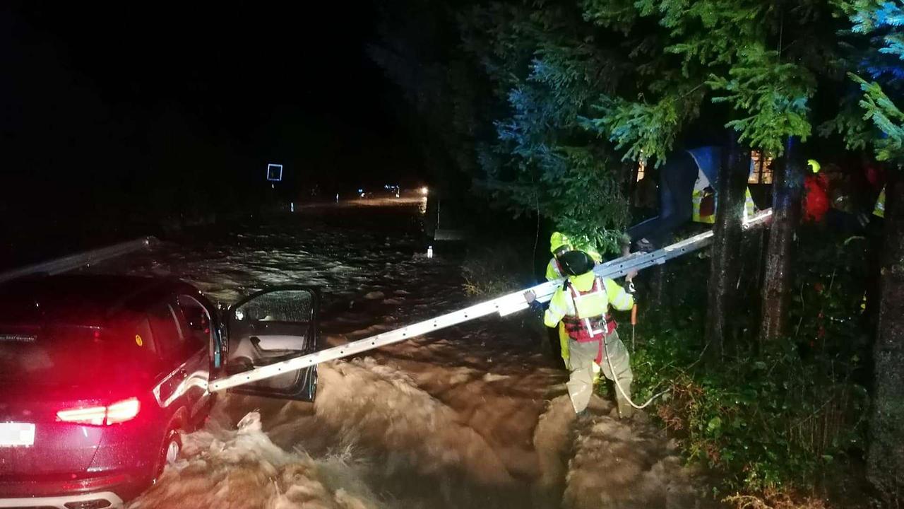 Ein Auto auf einer überfluteten Straße bei Nacht. Die Beifahrertür ist offen, von der Seite strecken Feuerwehrleute eine lange Leiter zum Auto. Sie sind mit Seilen gesichert.