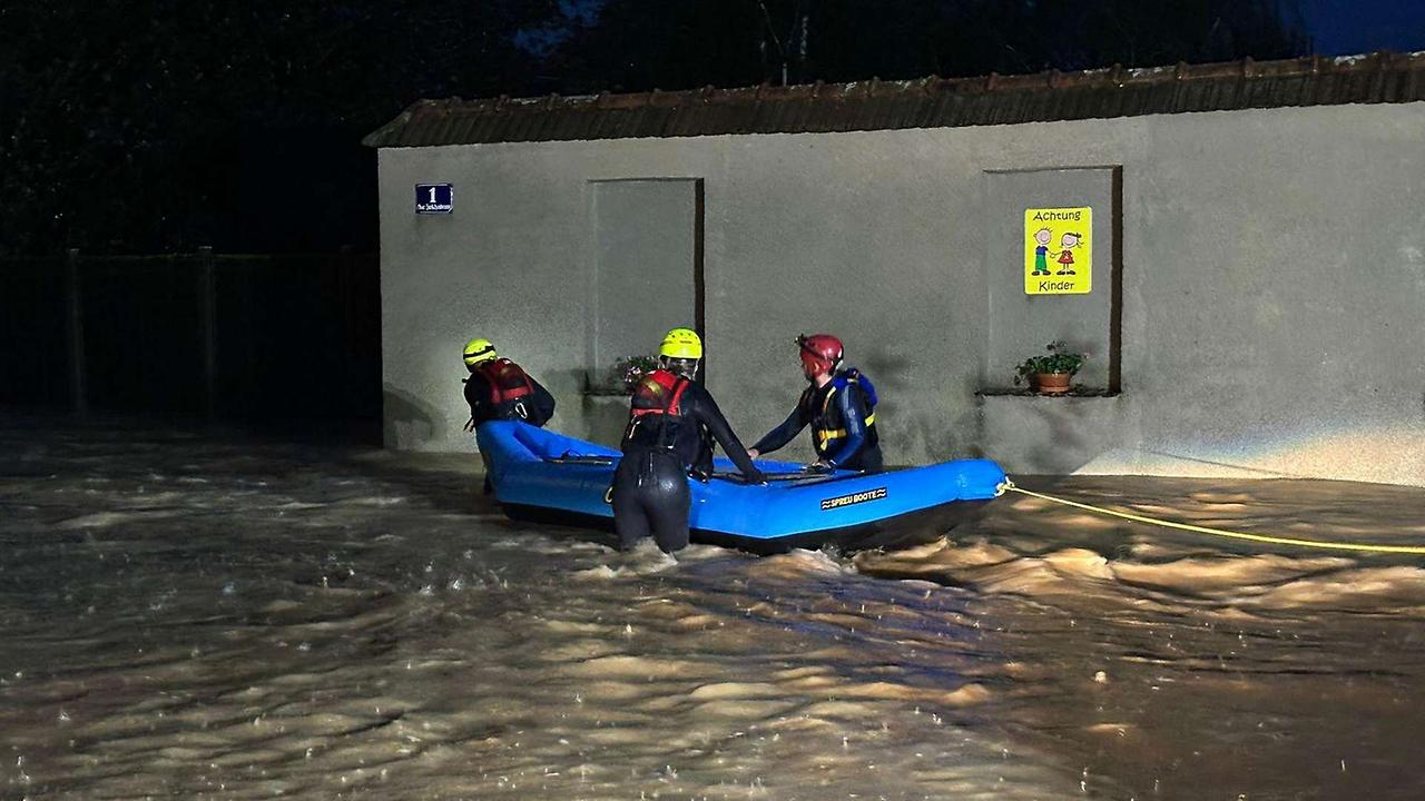 Drei Feuerwehrleute in Neoprenanzügen halten bewegen ein Schlauchboot in Richtung eines Hauses. Das Wasser geht ihnen bis zu den Oberschenkeln.