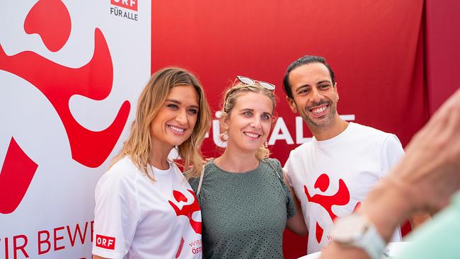 Eine junge Frau und ein Mann in „Wir bewegen Österreich“-T-Shirts posieren mit einer Frau für ein Foto. Alle lächeln, und im Hintergrund sieht man das rot-weiße Logo der Kampagne.