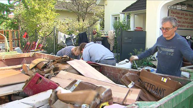 Eine Mulde steht auf der Straße vor Wohnhäusern. Sie ist fast bis zum Rand voll mit durchnässten Kartons und Holzplatten. Ein Mann mit ernstem Gesicht hat gerade etwas hineingeworfen.