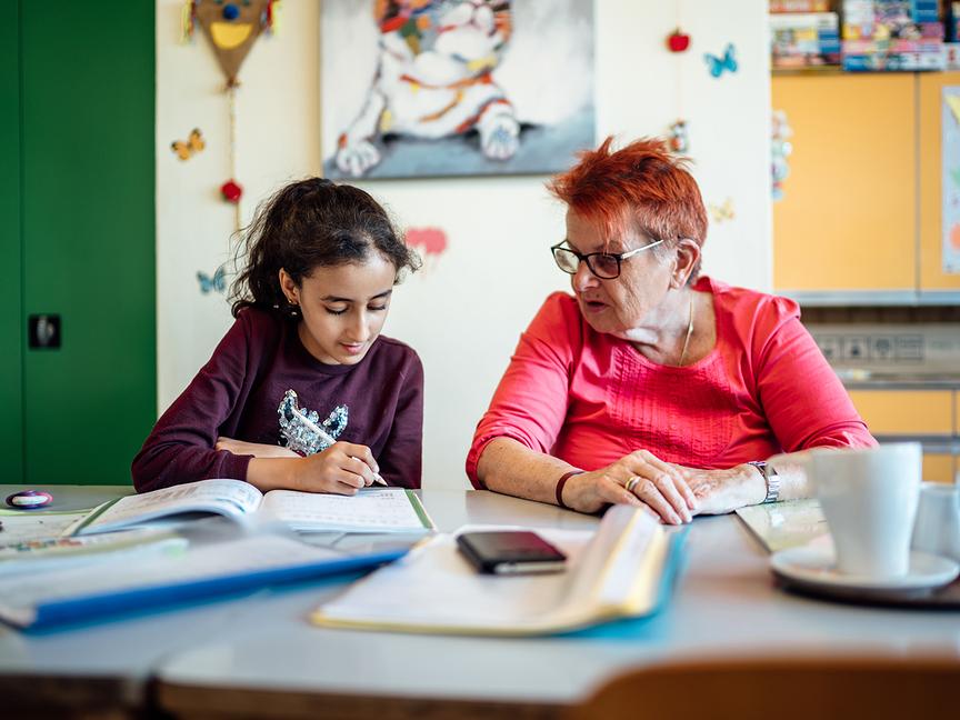 Ein etwa 10-jähriges Mädchen mit dunklen Haaren und eine ältere Dame mit rotgefärbten Haaren sitzen nebeneinander und blicken auf dasselbe Schulbuch. Im Hintergrund sind bunte Kästen und Bilder zu sehen, im Vordergrund liegen Schnellhefter und ein Notizbuch.