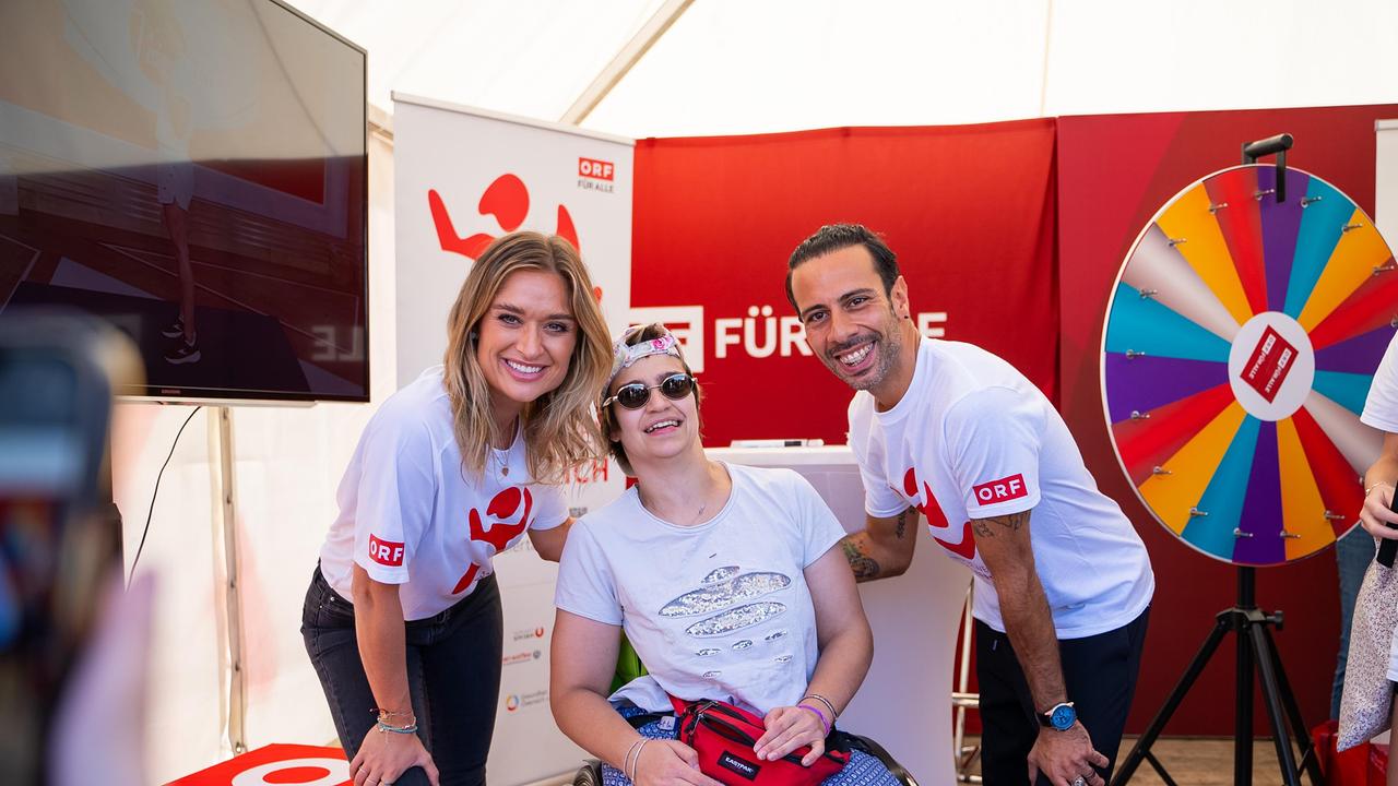 Eine junge Frau und ein Mann in "Wir bewegen Österreich"-T-Shirts posieren mit einer Person im Rollstuhl für ein Foto. Im Hintergrund ist ein buntes Glücksrad zu sehen, und alle Personen lächeln in die Kamera.