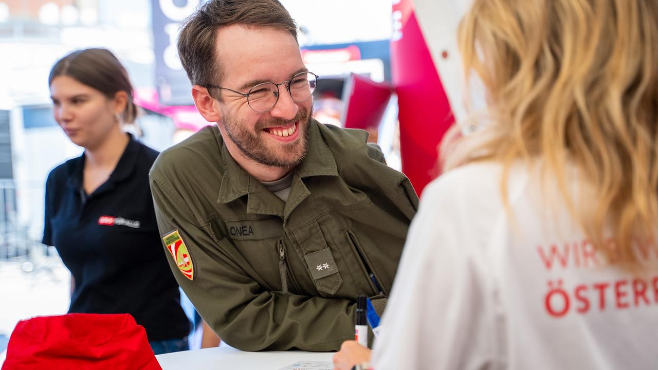 Ein Mann in Militäruniform lehnt sich lächelnd über einen Tisch und unterhält sich mit einer Person, die ein „Wir bewegen Österreich“-T-Shirt trägt. Im Hintergrund ist eine weitere Person in einem ORF-T-Shirt zu sehen.