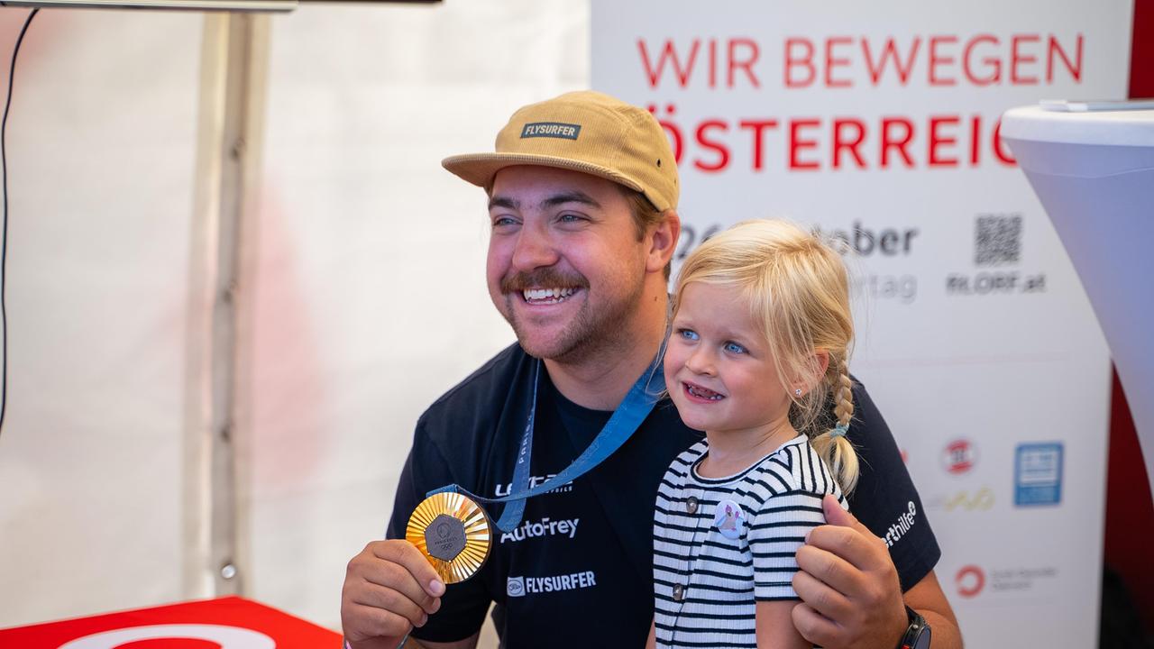 Ein Athlet mit einem breiten Lächeln und einer Medaille posiert mit einem kleinen Mädchen vor einem „Wir bewegen Österreich“-Banner. Beide schauen glücklich in die Kamera.