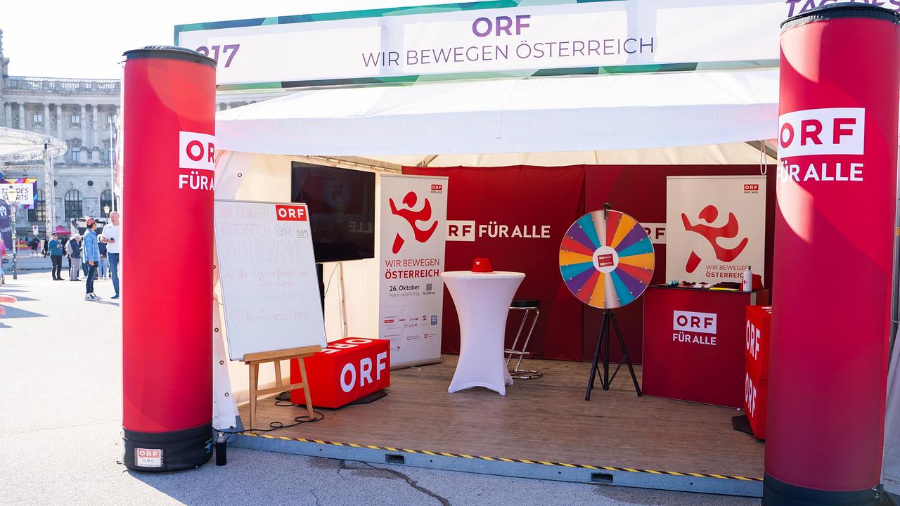 Ein ORF-Stand mit dem „Wir bewegen Österreich“-Logo und dem ORF-Logo. Der Stand hat eine rot-weiße Farbgebung. Ein Glücksrad und ein Tisch mit ORF-Branding sind zu erkennen.