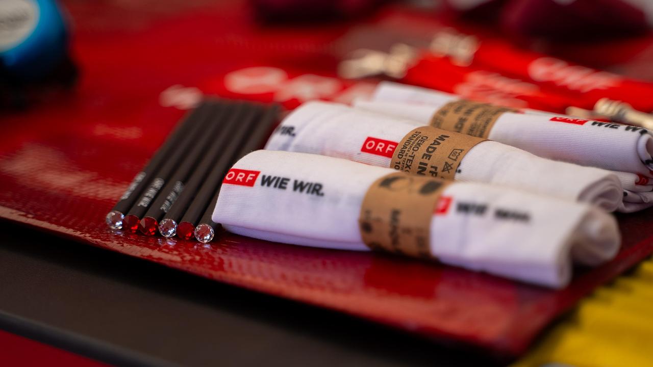 Nahaufnahme von Werbeartikeln, darunter gefaltete Bandanas mit dem ORF „Wie wir“-Logo und gebrandete Stifte, die auf einer roten Unterlage liegen.