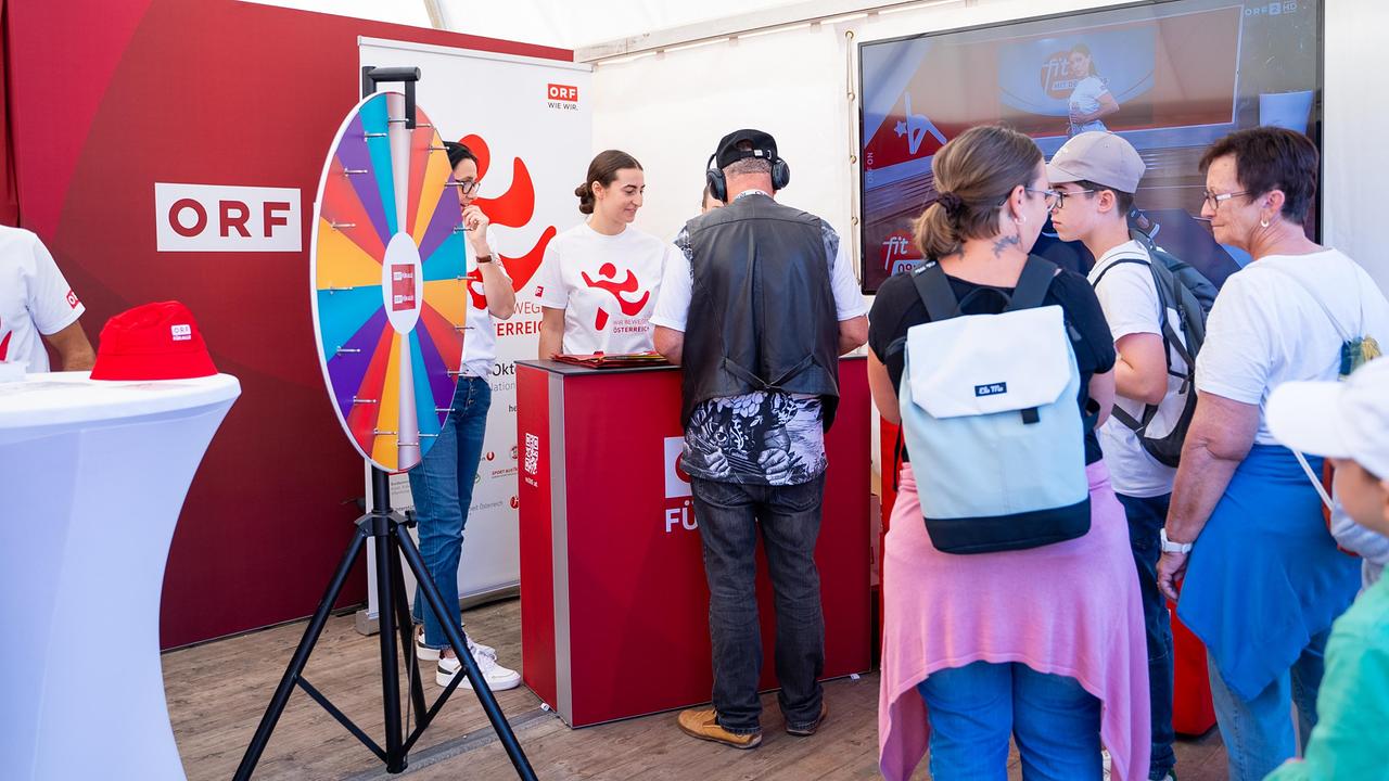 Im ORF-Stand. Ein buntes Glücksrad und ein Tisch mit ORF-Branding sind zu sehen. Mehrere Besucher*innen stehen am Stand und warten auf das Glücksrad. Im Hintergrund ein TV-Bildschirm mit einem Ausschnitt aus einer Mitturn-Sendung.